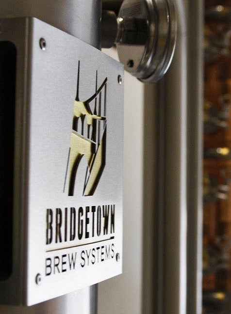 Bridgetown Brew Systems logo on a metal surface with brewing equipment in the background